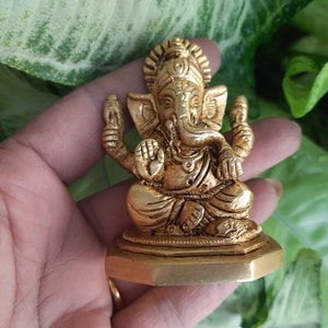 May include: A gold-coloured statue of the Hindu deity Ganesha, seated in a meditative pose. The statue has intricate details, including the elephant head, multiple arms, and ornate jewellery. The base is a hexagonal shape.