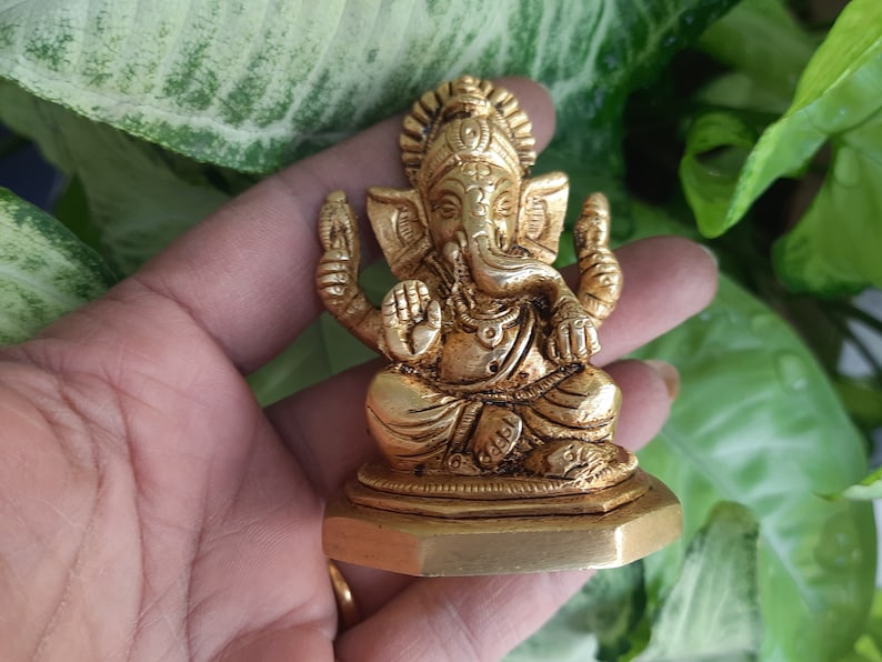 May include: A gold-coloured statue of the Hindu deity Ganesha, seated in a meditative pose. The statue has intricate details, including the elephant head, multiple arms, and ornate jewellery. The base is a hexagonal shape.