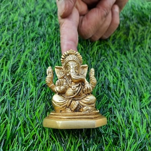May include: A gold-coloured statue of the Hindu deity Ganesha, seated on a small base. The statue is highly detailed, with intricate carvings. The background is artificial green grass, and a finger is pointing at the statue.