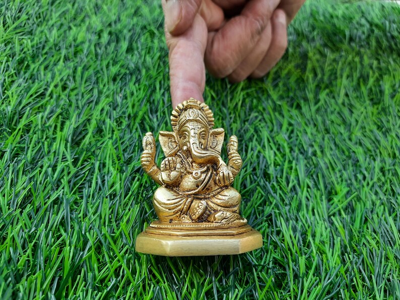 May include: A gold-coloured statue of the Hindu deity Ganesha, seated on a small base. The statue is highly detailed, with intricate carvings. The background is artificial green grass, and a finger is pointing at the statue.