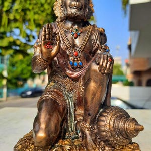 Puede incluir: Estatua de bronce de Hanuman, una deidad hindú, en posición de rodillas. La estatua está adornada con joyas coloridas y detalles intrincados. La figura tiene una cara de mono y está sentada sobre una base texturizada, con una caracola cerca.