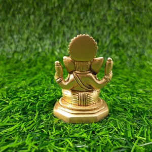 May include: A gold-coloured statue of Ganesha, the Hindu deity, seen from the rear. It is placed on a bed of bright green artificial grass. The statue features a detailed crown and multiple arms.