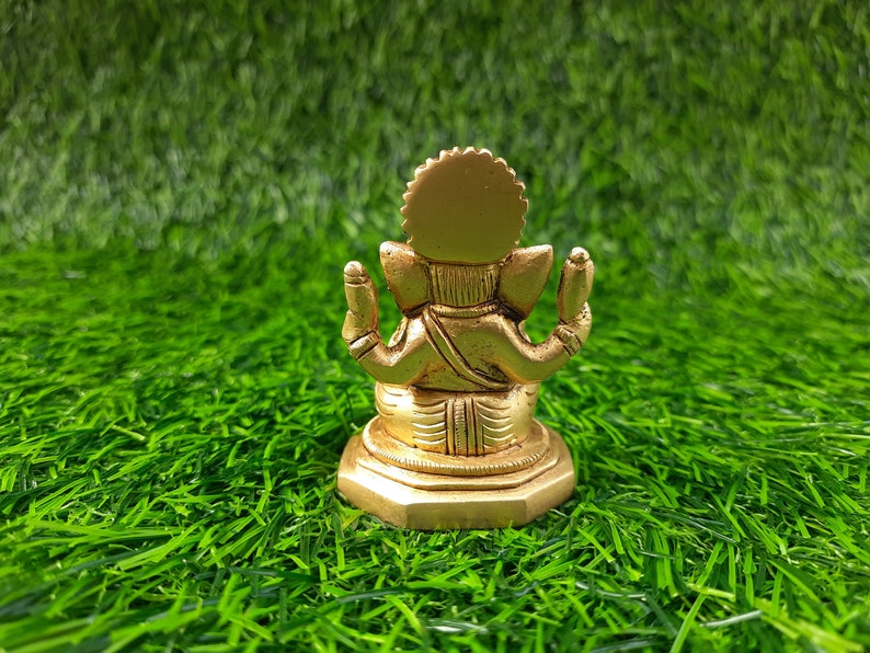 May include: A gold-coloured statue of Ganesha, the Hindu deity, seen from the rear. It is placed on a bed of bright green artificial grass. The statue features a detailed crown and multiple arms.