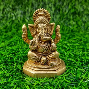 May include: A gold-coloured brass statue of Ganesha, the Hindu deity, in a seated position. The sculpture has detailed features, including an elephant head, multiple arms, and decorative jewellery. The statue is placed on a bed of green artificial grass.