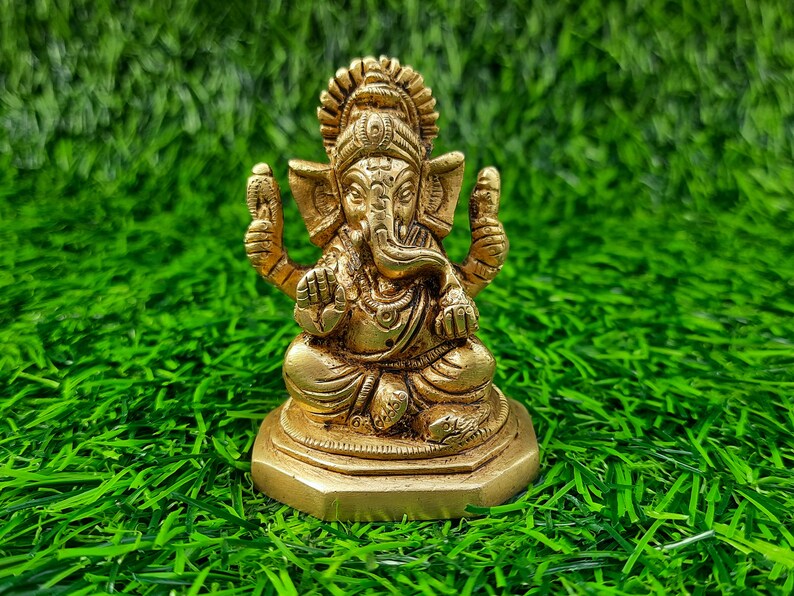 May include: A gold-coloured brass statue of Ganesha, the Hindu deity, in a seated position. The sculpture has detailed features, including an elephant head, multiple arms, and decorative jewellery. The statue is placed on a bed of green artificial grass.