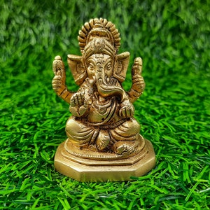 May include: A gold-coloured statue of Ganesha, the Hindu deity, in a seated posture. The statue features detailed carvings, including a large head, elephant ears, and multiple arms. It is placed on a hexagonal base against a background of green artificial grass.