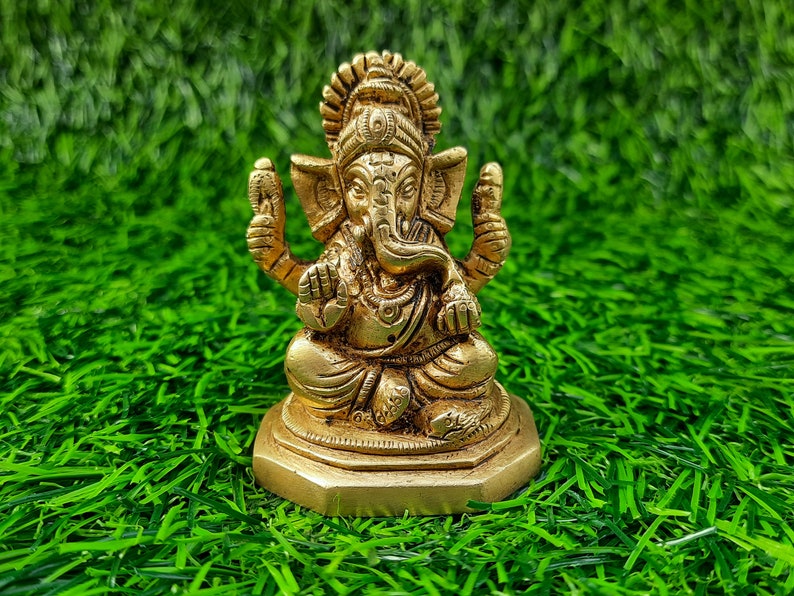 May include: A gold-coloured statue of Ganesha, the Hindu deity, in a seated posture. The statue features detailed carvings, including a large head, elephant ears, and multiple arms. It is placed on a hexagonal base against a background of green artificial grass.