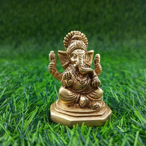 May include: A small, gold-coloured statue of the Hindu deity Ganesha, seated with multiple arms and an elephant head. The statue is on a square base and set against a backdrop of green artificial grass.