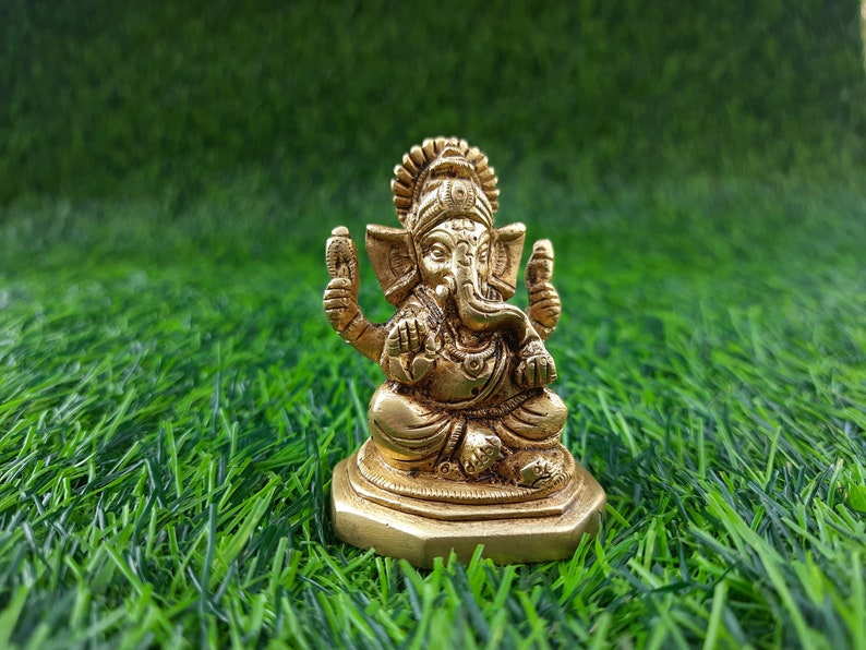 May include: A small, gold-coloured statue of the Hindu deity Ganesha, seated with multiple arms and an elephant head. The statue is on a square base and set against a backdrop of green artificial grass.
