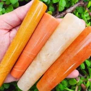 Op de afbeelding: Vier gepolijste stenen pijpen in tinten oranje, geel en crème worden getoond. De pijpen zijn glad en cilindrisch, met een taps toelopende vorm. De achtergrond is een levendig groen met kleine bladeren.