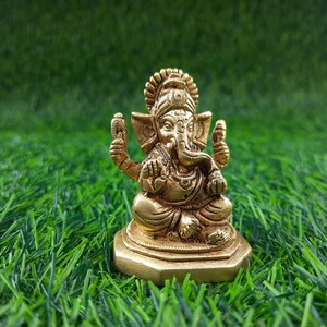 May include: A small, gold-coloured statue of the Hindu deity Ganesha, seated in a meditative pose. The statue has intricate details, including a large head, elephant ears, and multiple arms. It is set against a backdrop of green artificial grass.