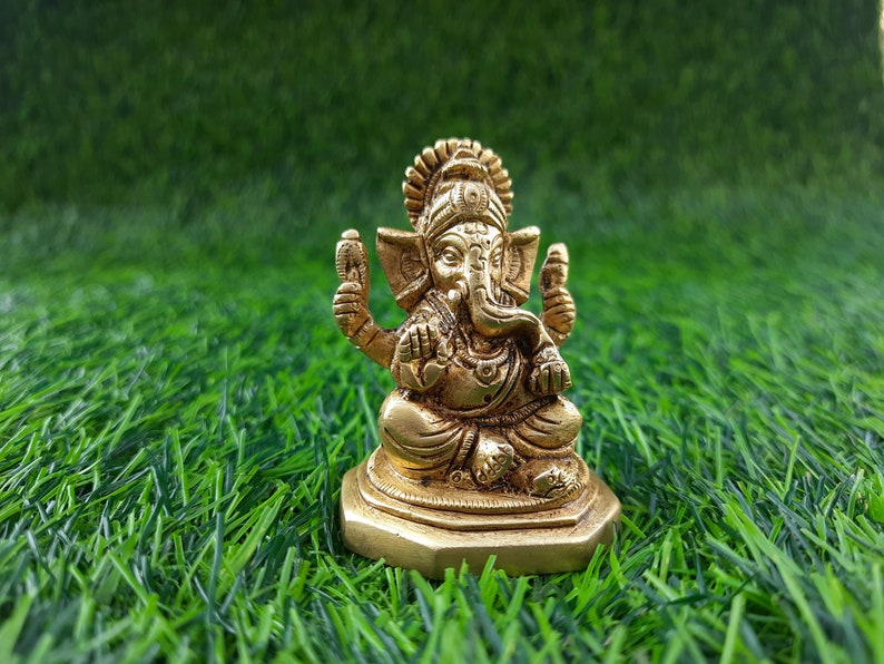 May include: A small, gold-coloured statue of the Hindu deity Ganesha, seated in a meditative pose. The statue has intricate details, including a large head, elephant ears, and multiple arms. It is set against a backdrop of green artificial grass.