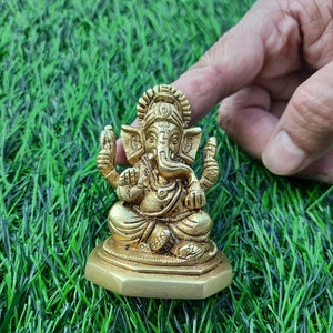 May include: A golden brass statue of the Hindu deity Ganesha, seated in a meditative pose. The statue features intricate details and is placed on a hexagonal base. The background is artificial green grass.