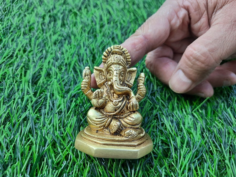 May include: A golden brass statue of the Hindu deity Ganesha, seated in a meditative pose. The statue features intricate details and is placed on a hexagonal base. The background is artificial green grass.