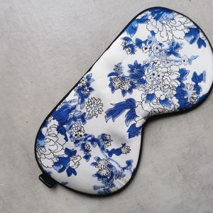 May include: A white sleep mask with a blue and white floral pattern. The mask has a black elastic band.