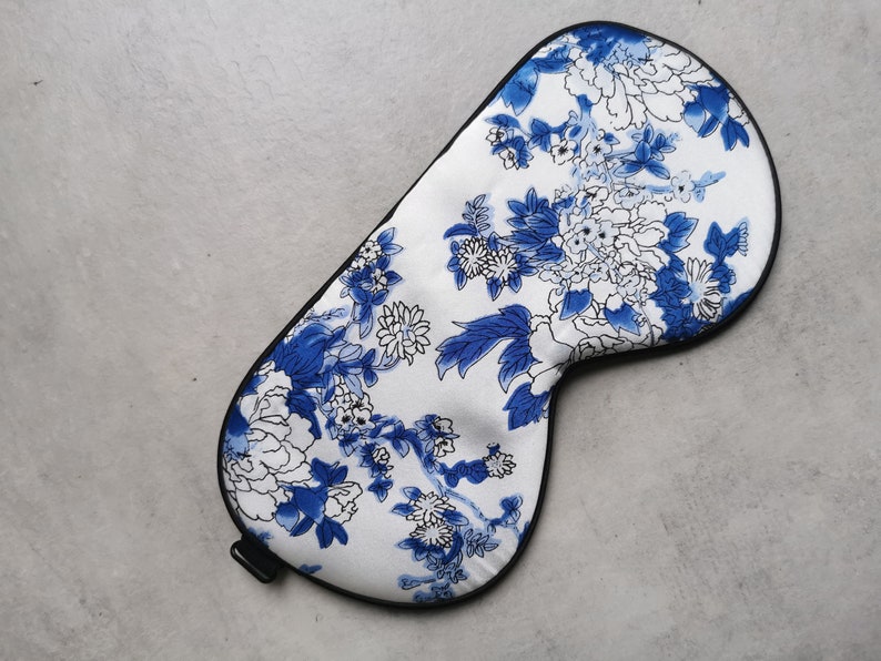 May include: A white sleep mask with a blue and white floral pattern. The mask has a black elastic band.