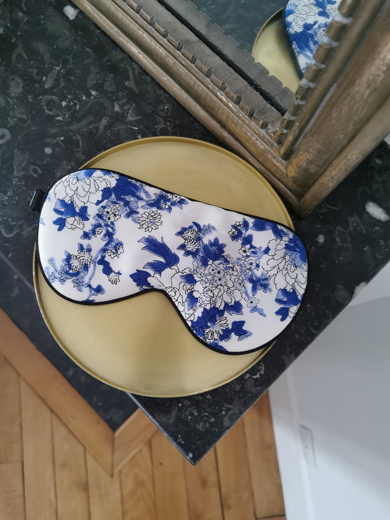 May include: A white sleep mask with a blue and white floral pattern. The mask has a black elastic band.