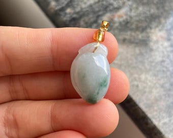 Puede incluir: Un colgante de jade con una fianza dorada. El colgante es de color verde claro con vetas verdes más oscuras. El colgante es liso y pulido, con forma de lágrima.