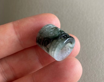 Puede incluir: Una pequeña cuenta de jade cilíndrica de color verde y blanco con un patrón de remolino.