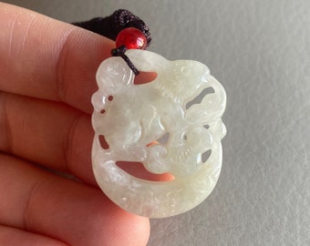 May include: A white jade pendant with a carved design of a dragon. The pendant is attached to a dark brown cord with a red bead.