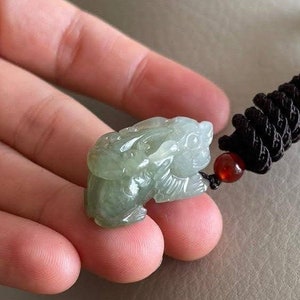 May include: A jade green pendant carved in the shape of a mythical creature, attached to a black braided cord with red beads.