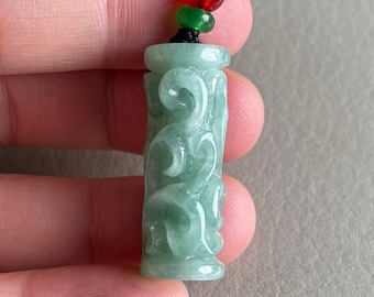 Puede incluir: Un colgante de jade verde claro con intrincados diseños tallados. El colgante cilíndrico está suspendido de un cordón negro con detalles rojos, verdes y dorados. El jade es translúcido.