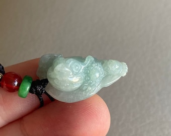 May include: A jade green pendant carved in the shape of a frog, with intricate details. The pendant is attached to a black cord with red and green beads.