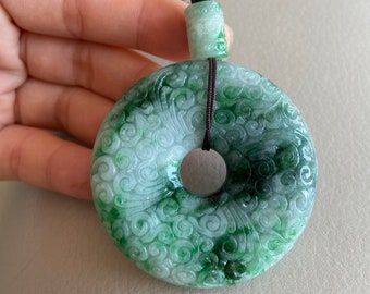 May include: A green jade pendant with a carved swirl design. The pendant is attached to a brown cord with a green bead.