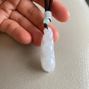 May include: A white jade pendant necklace with a string of three round beads. The pendant is shaped like a pea pod and is attached to a brown cord.