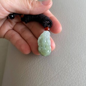 May include: A green jade pendant carved with a detailed design, hanging from a brown cord with red beads.