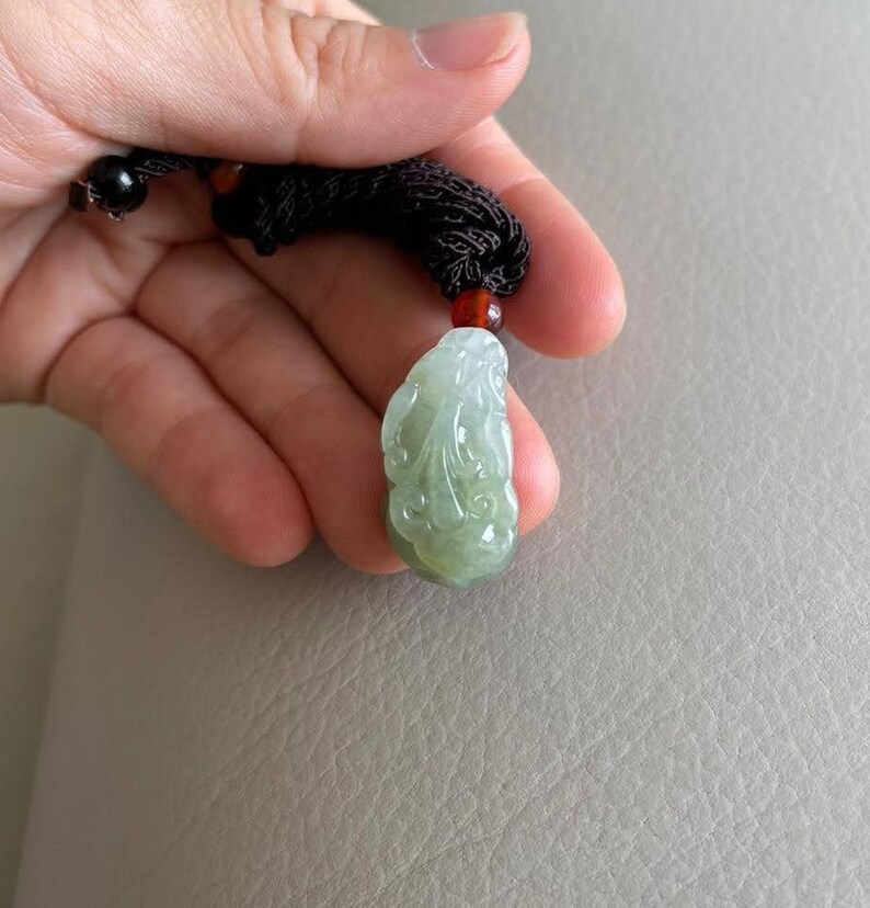 May include: A green jade pendant carved with a detailed design, hanging from a brown cord with red beads.