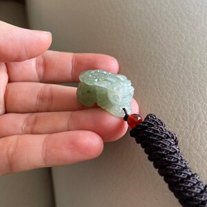 May include: A green jade pendant carved in the shape of a mythical creature, hanging from a brown cord with a red bead.