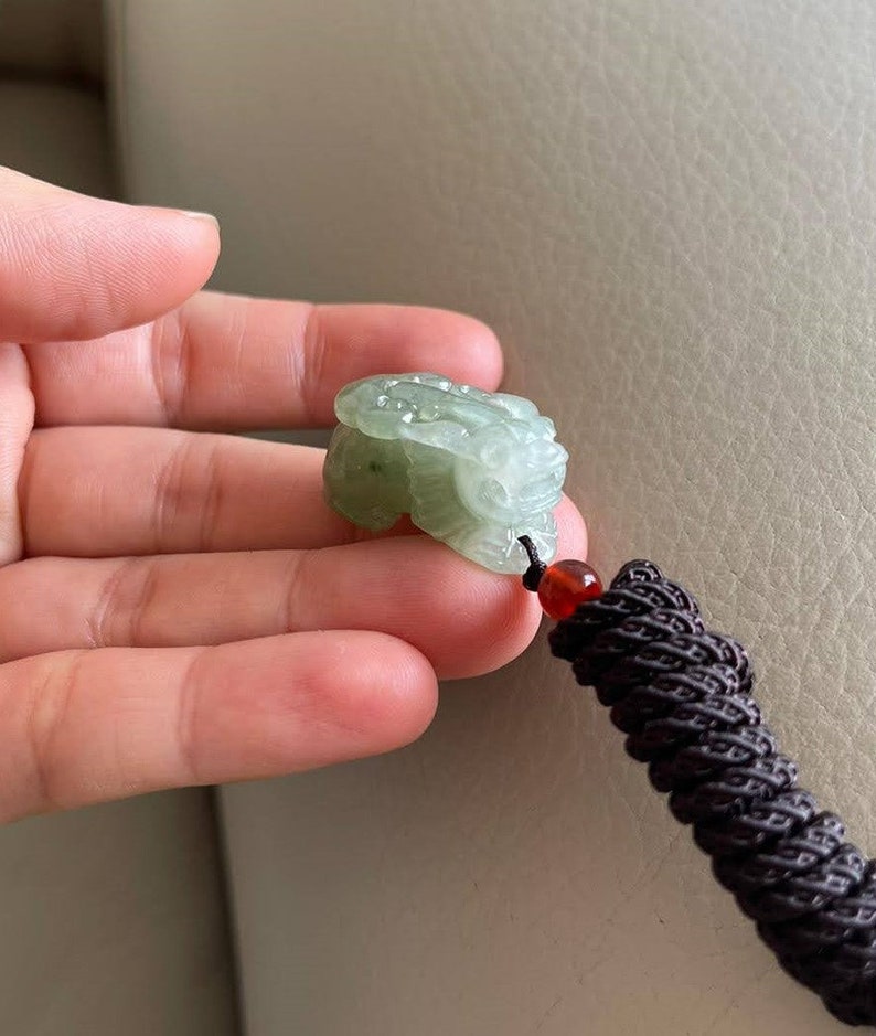 May include: A green jade pendant carved in the shape of a mythical creature, hanging from a brown cord with a red bead.