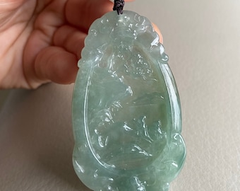 May include: A green jade pendant with a carved design of a landscape scene. The pendant is attached to a brown cord with gold accents.
