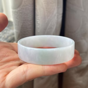 May include: A light green and white jade bangle bracelet. The smooth, polished surface reflects light, showcasing the natural variations in color. The bracelet is a solid, circular band, perfect for everyday wear.