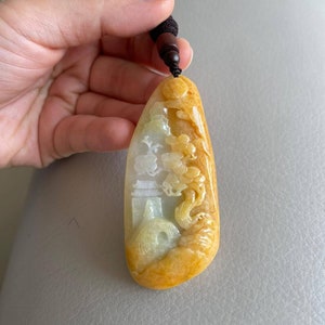 May include: A yellow jade pendant with a carved design of a dragon and a pagoda. The pendant is attached to a brown cord.