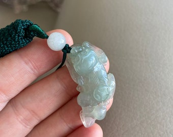 May include: A translucent jade pendant carved in the shape of a mythical creature. The pendant is light green with subtle variations in color. It is attached to a dark green knotted cord with a white bead.