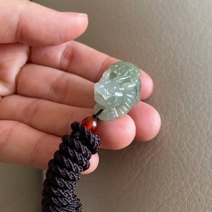 May include: A jade green dragon pendant with intricate details, hanging from a brown braided cord with red and orange beads.