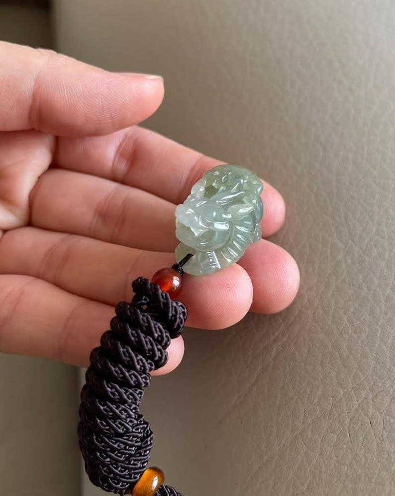 May include: A jade green dragon pendant with intricate details, hanging from a brown braided cord with red and orange beads.