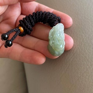 May include: A jade pendant with a carved design, attached to a brown braided cord with black and amber beads.