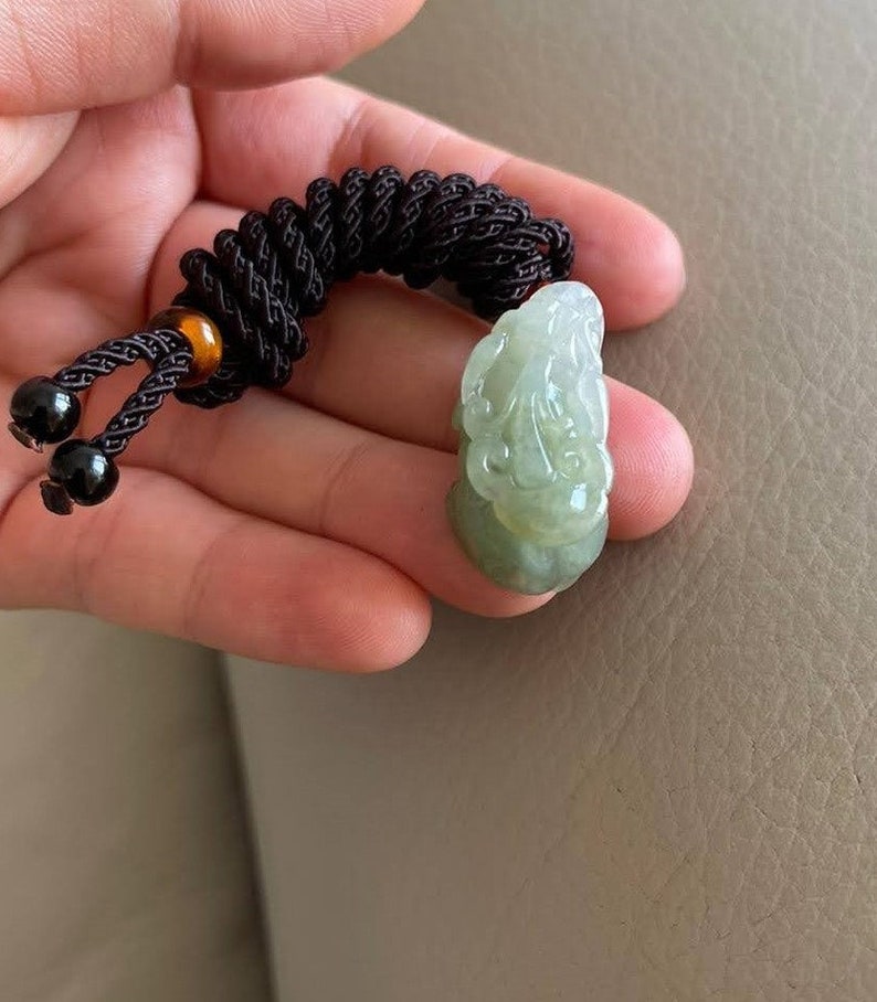 May include: A jade pendant with a carved design, attached to a brown braided cord with black and amber beads.