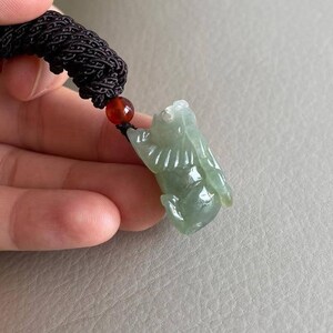 May include: A jade green pendant carved in the shape of a mythical creature, hanging from a black cord with a red bead.