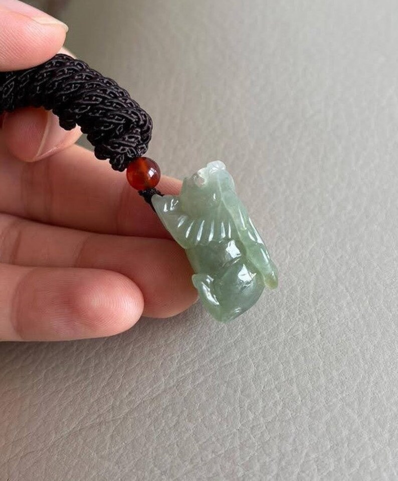 May include: A jade green pendant carved in the shape of a mythical creature, hanging from a black cord with a red bead.