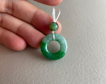 May include: A green jade pendant with a round shape and a hole in the center. The pendant is attached to a white string.