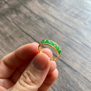 May include: A gold band ring with five green jade stones set in a row. The ring is on a wooden surface.