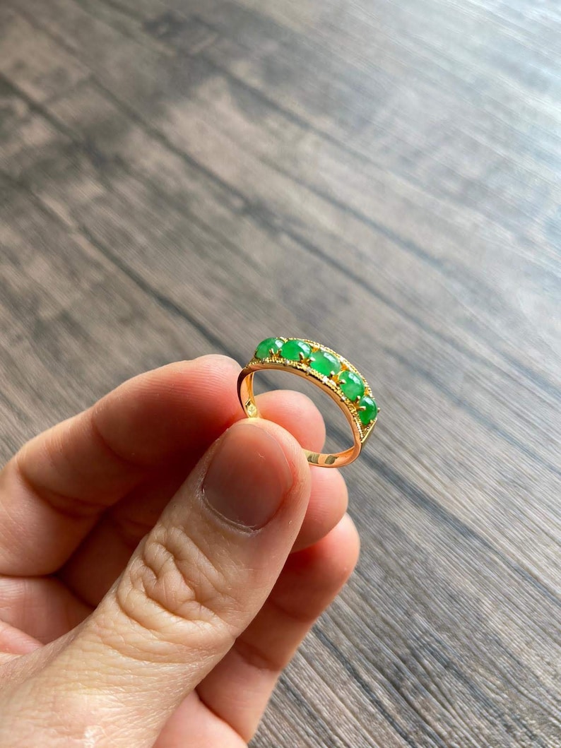 May include: A gold band ring with five green jade stones set in a row. The ring is on a wooden surface.