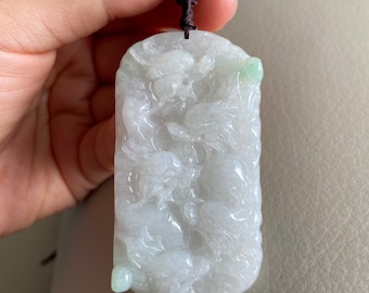 May include: A white jade pendant with intricate carvings, featuring a light green jade accent. The pendant is attached to a brown cord.
