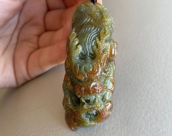 Op de afbeelding: Een groene en bruine jade hanger gegraveerd in de vorm van een draak. De hanger hangt aan een bruin koord met gouden accenten.