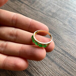 May include: A gold band ring with a row of green gemstones. The ring is on a person's hand.
