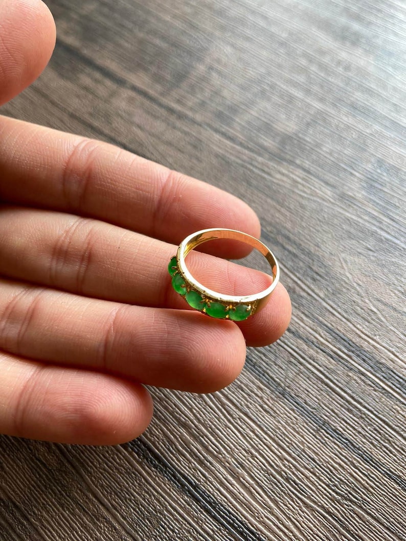 May include: A gold band ring with a row of green gemstones. The ring is on a person's hand.